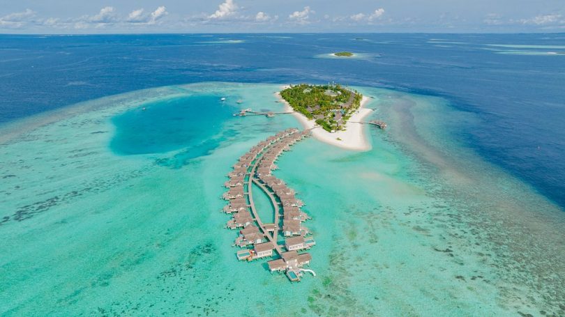 Luxury Escapes Aerial view of a tropical island with overwater bungalows surrounded by clear blue water.