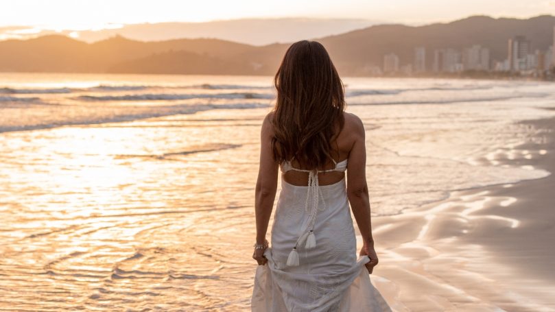 Freedom Escapes Woman in a white dress walking barefoot along a beach at sunset.