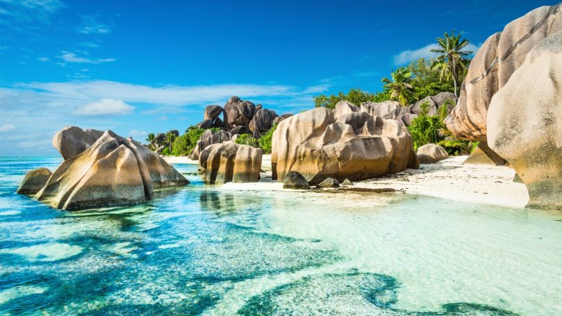 Signature Seychelles Idyllic beach scene with large rocks, clear water, and lush greenery under a blue sky.